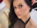 HannaRobertss has long black hair, brown eyes, and pink lips, wearing a black mesh top with a halter neck, and silver earrings. She's looking at the camera.