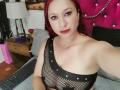 MonicaLew with red hair, black mesh top, and red lipstick, showing her bare tits, a slight smile, and holding a keyboard. Background includes a pink tufted headboard.