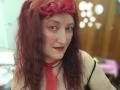 Sabrhina with red curly hair, pink face makeup, red bow, red lipstick, and red necktie, showing her boobs in a black top, smiling at the camera.
