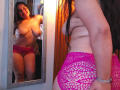 SammyTitss is topless with large boobs, wearing pink mesh panties, standing in front of a mirror. Her reflection shows a smile, brown hair, and a wooden door.