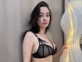 JuleeKasprzak with pale skin, dark wavy hair, wearing a black sheer bra with strappy details, standing next to a wavy white lamp, holding it gently.