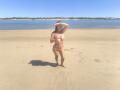 DosCalientes stands on a sandy beach, covering her bare tits with one hand, wearing an orange bikini, and shielding her eyes with the other, under a clear blue sky.