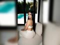 MiilaWx with long black hair, white bra, and white ass-hugging pants, sitting on a white couch, looking over her shoulder, by a blue pool, outdoors, sunny.