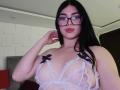IsabellaaRossi with long black hair, glasses, and red lipstick, wearing a white lace bra with black bows, showing her tits. She's touching her hair.