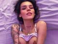LenaBranch with tattoos on her arm and chest, lying on a purple bed, wearing a white bra that exposes her tits, looking up with a slight smile.