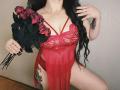Niferttiti with long black hair, wearing red lace lingerie, holding a bouquet of red roses, showing tattoos on thigh, black nails, and a gold necklace.