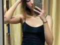 HeikeCarchi with medium tits in a black spaghetti strap top, taking a mirror selfie with her phone, blonde hair, right arm raised, yellow curtains in the background.