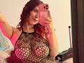 ValeryScot with curly red hair, wearing a black fishnet top, showing large tits, holding a pink phone, pink lighting, mirror selfie, bed with white and pink pillows.