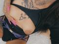 DenyChantal's black top exposing her large, tan tits with a black bra, purple shorts, pink nails, tattoos, and a hand covering her nipple. Her long black hair is visible.