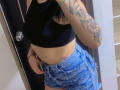 Kkatalina with a black sports bra, showing her tits, wearing ripped blue denim shorts, tattoo on right arm, holding phone, and a red bracelet, in a selfie.