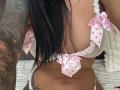 AndraD with large tits in white polka dot bra with pink bows, showing tattooed arm and black hair, wearing white panties, in a slightly angled selfie.