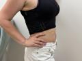 Tilliana's side profile showing her black sports bra with cutout sides, small to medium tits, and white jeans. She's standing with one hand on her hip.