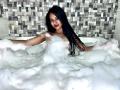 EstrellaXHot with long black hair, red lipstick, and visible tits, sitting in a bubbly white bath, arms out, surrounded by foam, in a tiled bathroom.