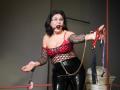 KarlaHartley with black hair, glasses, red bra, fishnet top, black latex pants, ball gag, holding red and purple restraints, pink cuffs, chain, and white statue.