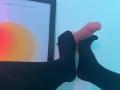 Les pieds d'IrisBlair, portant des chaussettes noires, agrippent un pénis grand, érigé et non circoncis contre un mur bleu clair. Les orteils d'IrisBlair sont enroulés autour de la base..
