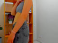 Steisii with gray bra and high-waisted denim skirt, side profile, arm raised, showing her ass and slim waist, orange bookshelf with plants and bottle in the background.
