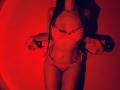 Tisia with long dark hair, wearing red lace lingerie, holds the sides of her panties, showing her small boobs and trimmed pussy, under red lighting.