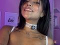 MajoJones mit glänzenden Lippen, dunklem Haar, einer weißen Spitzenkette, einem schwarzen 'P'-Anhänger, Tattoos auf der Brust, lächelnd, in violettem Licht, zeigt ihre Brüste, eine Halskette mit einem Mond-Anhänger, in einem Selfie..