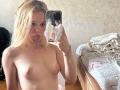 DominicaChambley, topless, blonde, small tits, sticking out tongue, holding phone, mirror selfie, phone case with white pop-sickle sticker, bed with gray and red sheets in background.