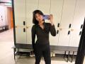 HeikeCarchi with dark hair, pouty lips, black zip-up, and black pants, taking a mirror selfie, holding a blue phone, standing in front of white lockers with black handles.