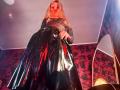 FiorellaNox with long blonde hair, wearing a shiny black vinyl skirt and sparkly silver top, looking down, her red lips and dark makeup standing out. Low angle shot.