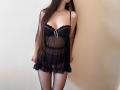 Tisia heeft lang bruin haar, draagt een zwarte, doorschijnende babydoll met een witte strik en zwarte netpanty's. Haar borsten zijn zichtbaar door de doorschijnende stof..