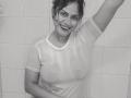 Black and white photo of IvannaDuran with wet shirt, showing her tits through the fabric, smiling, arm raised, curly dark hair, white tiled background, right hand on her belly.
