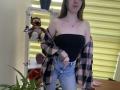Nesie with bare tits in a black strapless top, blue jeans, and a plaid shirt off her shoulders, standing in a room with a tiger stuffed animal.