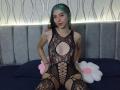 AbbyHillsI with blue-green hair, wearing a black lace bodysuit with large cutouts, exposing her tits and pussy, sitting on a bed with a gray headboard.