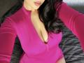 Olieri has big tits in a pink zip-up bodysuit, showing a deep cleavage with her left breast slightly exposed, dark wavy hair, and pink lips.