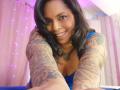 Chili with tattoos on arms, smiling, wearing blue top, reaching towards camera, dark wavy hair, purple lights in background, cute, brown eyes, small nose, visible necklace, medium tits.