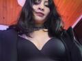 IssisDiosa with black wavy hair, wearing a black choker and black bra, showing her tits in a low-angle shot, wooden ceiling in the background, slight pink light.