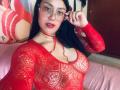 CristalZafiro lleva una blusa roja de malla transparente que deja ver sus pechos y un collar rojo. Tiene el pelo largo y negro, gafas y pintalabios rojo, y está ajustándose las gafas..