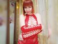 LaylaHottyX with red bob, wearing red lace-up dress and matching arm sleeves, smiling, arms crossed, showing her tits, standing in a room with fairy lights.