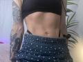 HotCaramelsTrio's midriff showing a pierced belly button, wearing a black crop top, studded blue jeans, and black straps, with tattooed arms and a black scrunchie.