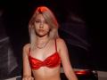 DataSley with pale skin, blonde hair, and red satin bra, wearing a silver chain necklace, looking serious, in a dimly lit setting, with red light highlighting her hair.