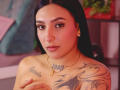 SophiieBlair with dark hair, brown eyes, nose ring, and gold earrings, topless, showing tattoos on chest and arm, covering her tits with one hand. Soft pink and orange lighting.